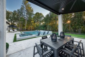 A modern outdoor patio from the White Oak outdoors portfolio features a dark gray dining table with eight chairs, two decorative lanterns, and a view of a spacious swimming pool with submerged lounge chairs. Beyond the pool are additional lounge chairs and a backdrop of tall trees under a clear sky.